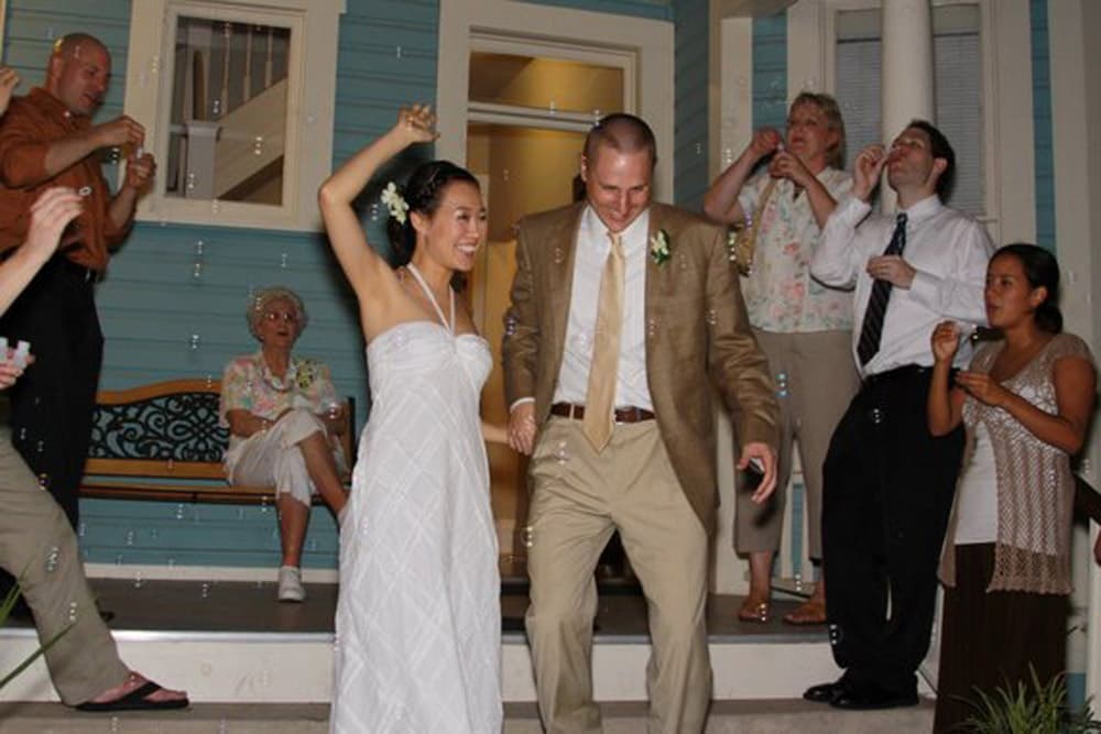 A bride and groom joyfully exit a house surrounded by family and friends celebrating with bubbles.