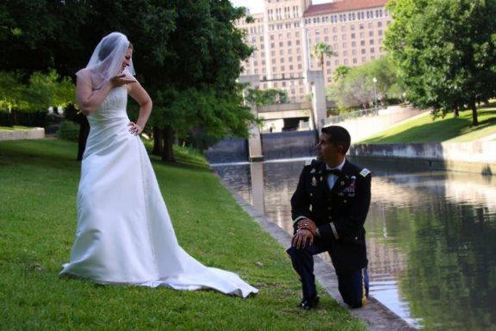 A bride in a white gown poses confidently by a river while a soldier kneels beside her.