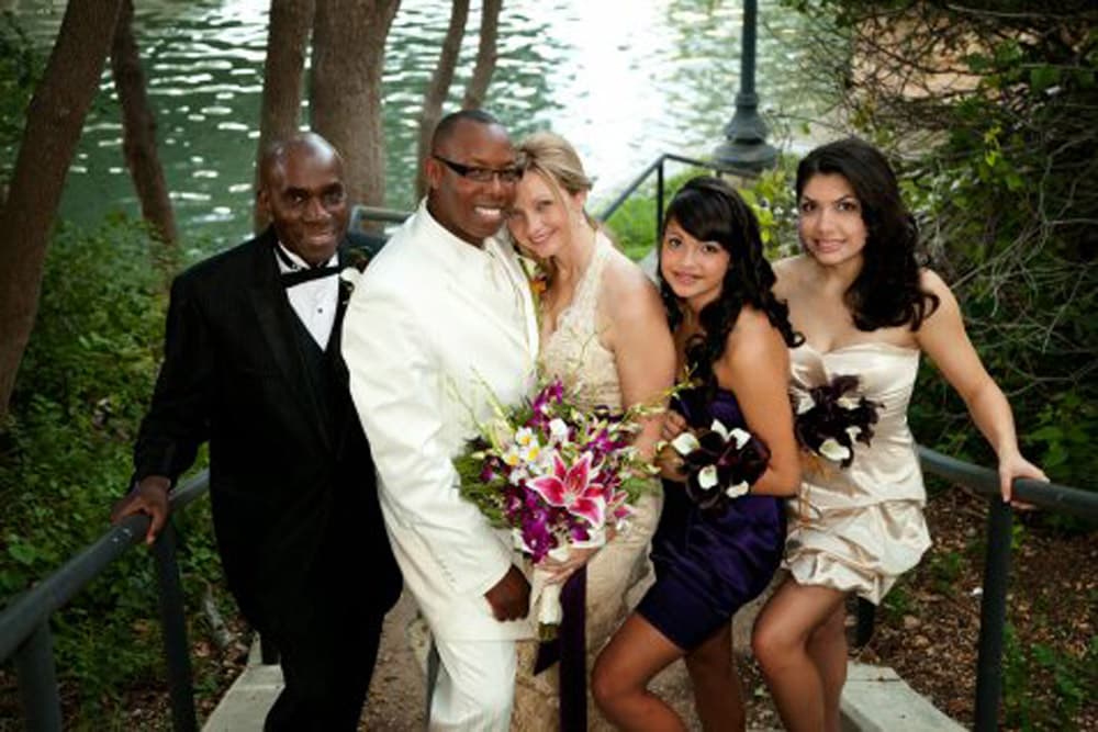 A group of five people poses together outdoors near a body of water, dressed in formal attire with floral arrangements.