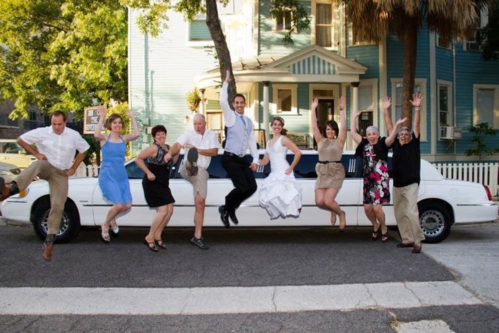 A group of joyful people, including a bride in a wedding dress, jump and pose in front of a limousine.