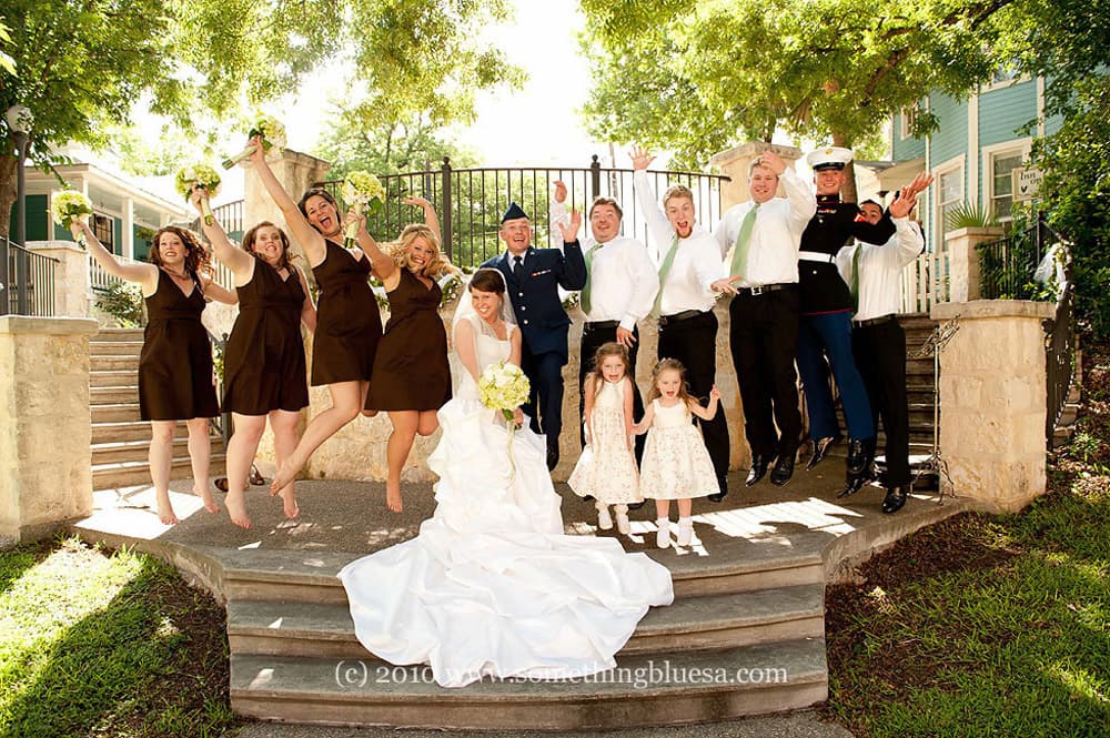 A joyful wedding party poses on steps, celebrating with arms raised and smiles.