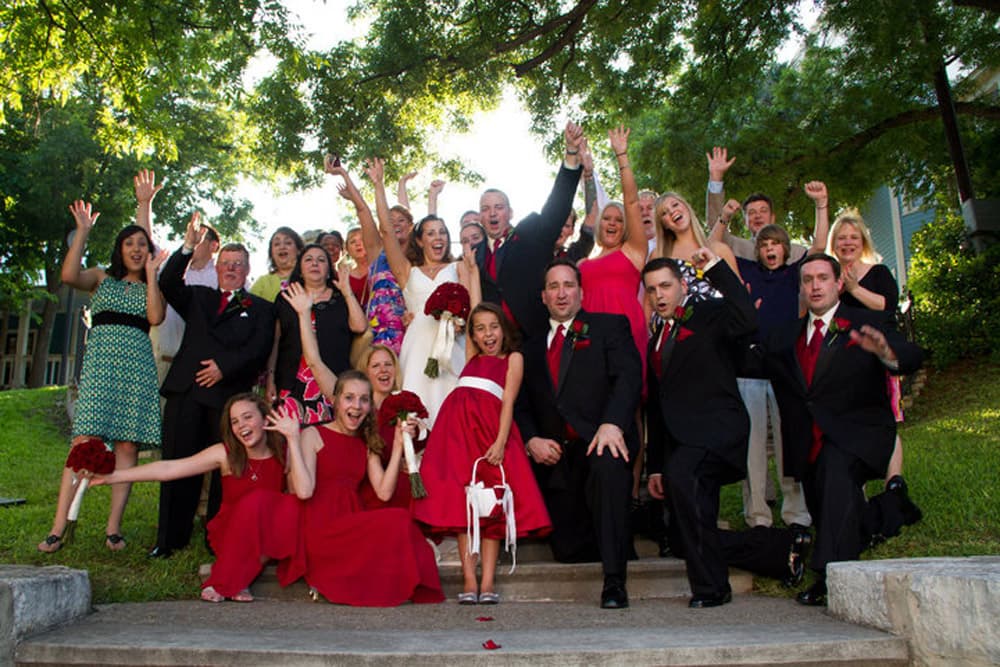 A large group of people in formal attire joyfully pose with raised hands outdoors.