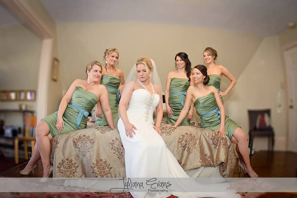 A bride in a white gown sits on a bed surrounded by five bridesmaids in green dresses.