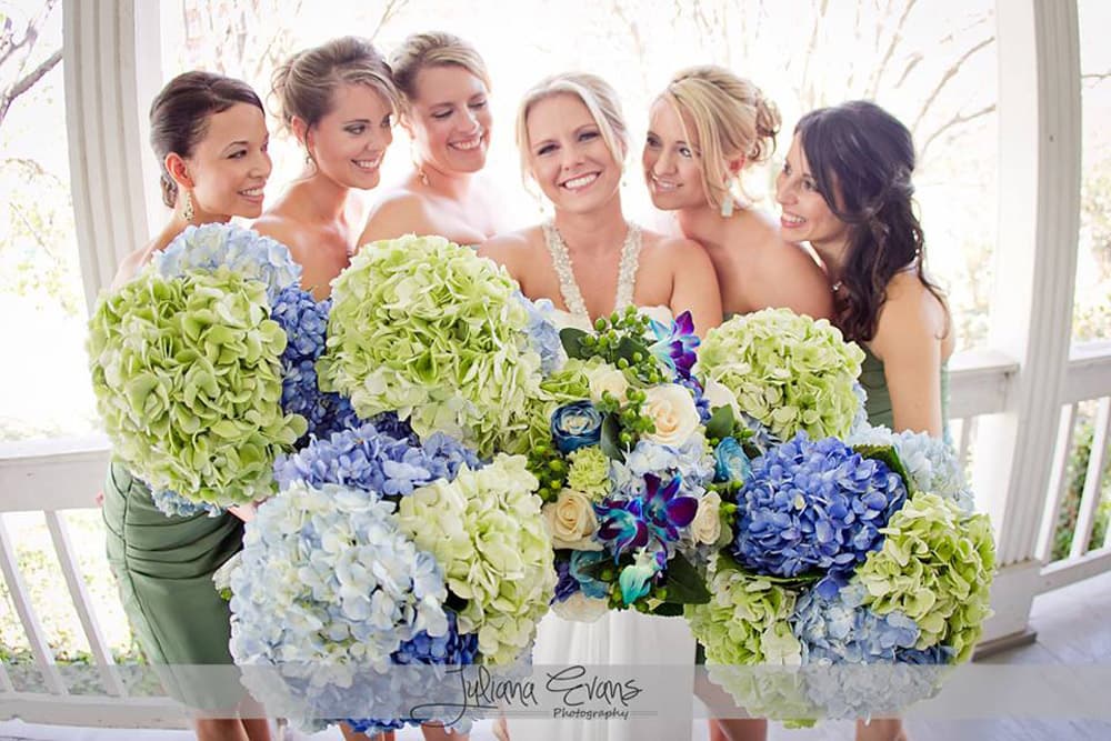 A smiling bride and her six bridesmaids pose together, holding large bouquets of blue and green flowers.
