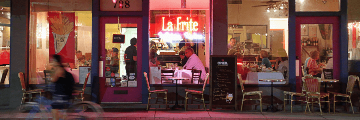 A warmly lit restaurant with diners visible through large windows, featuring a neon sign that reads "La Frite."