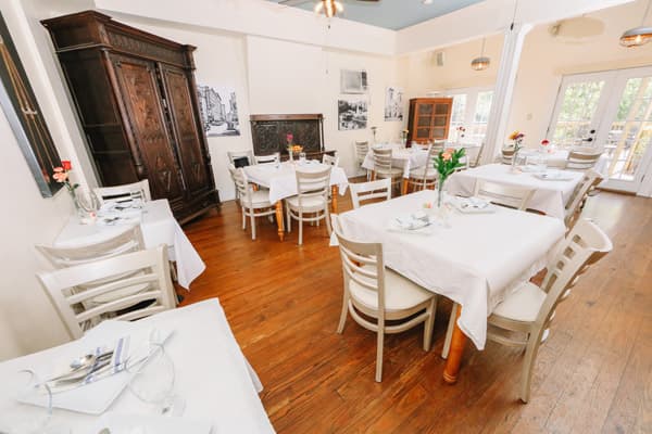 A cozy dining room with wooden floors, white tablecloths, and an antique cabinet, featuring several tables set for guests.