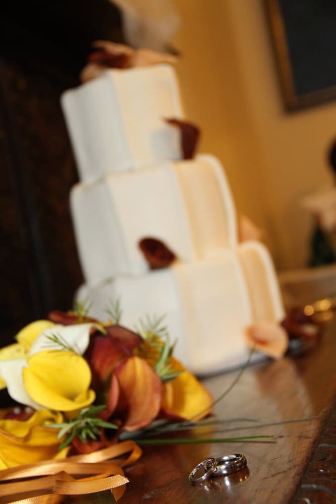 A three-tiered wedding cake is displayed alongside a bouquet of flowers and wedding rings on a wooden surface.