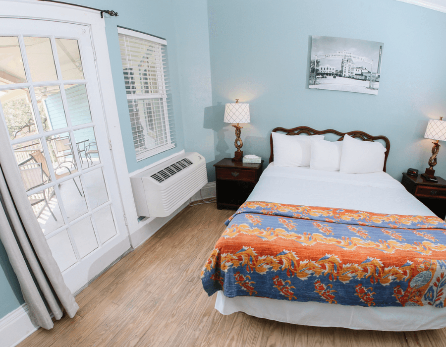 A bright bedroom featuring a double bed with a colorful blanket, two bedside lamps, and access to a balcony through French doors.