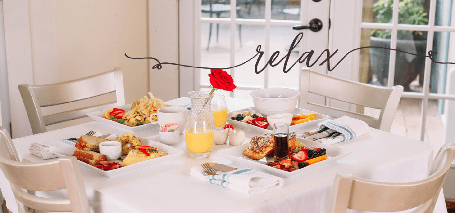 A beautifully arranged breakfast table with various dishes, a flower centerpiece, and the word "relax" elegantly scripted above.