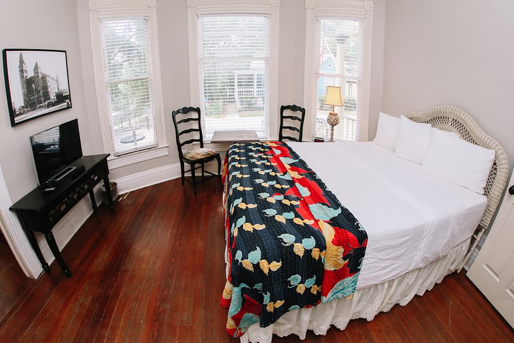 A cozy bedroom with a large bed, colorful quilt, wooden floor, and two windows allowing natural light.
