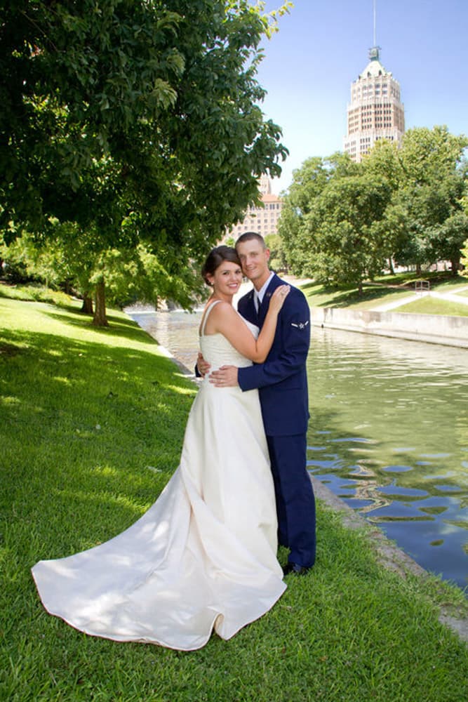 A bride in a white gown embraces a groom in military dress by a serene waterway surrounded by greenery.