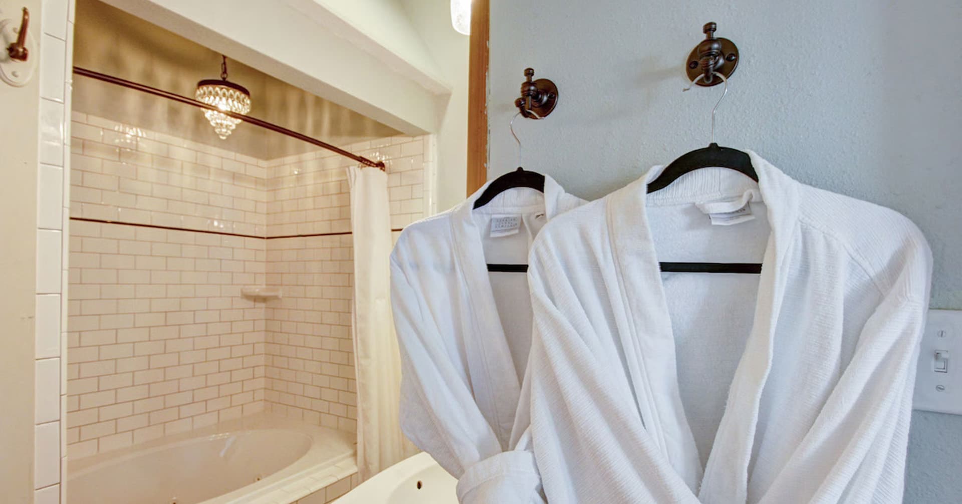 White bathrobes hang on hooks in a bathroom with a bathtub and tiled walls.