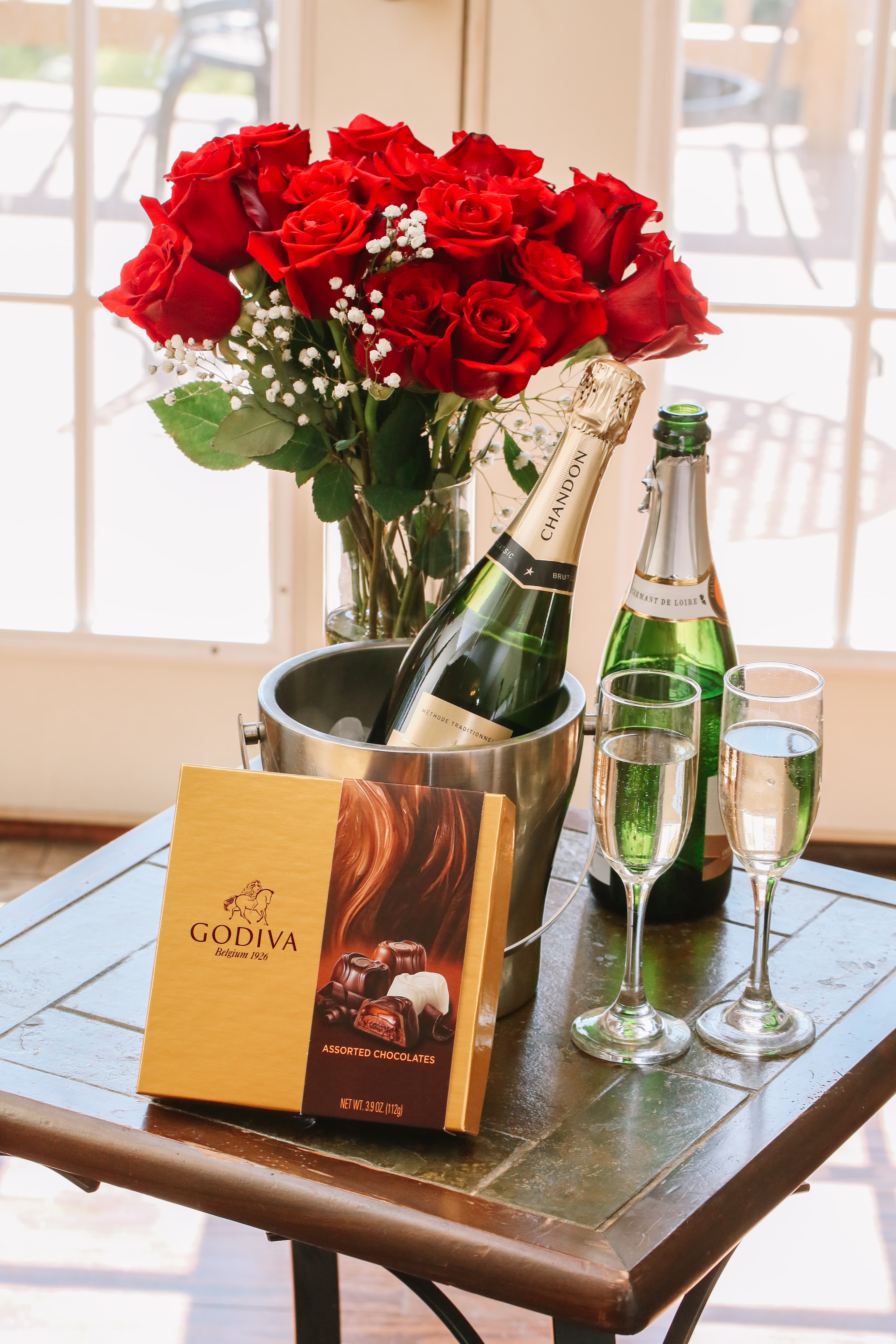 A bouquet of red roses, champagne bottles in a bucket, and a box of Godiva chocolates are arranged on a wooden table.