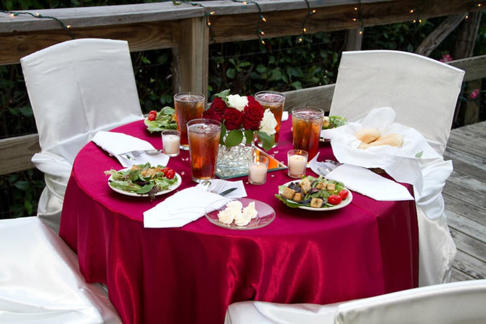 A elegantly set round table with a red tablecloth, adorned with salads, drinks, candles, and roses.