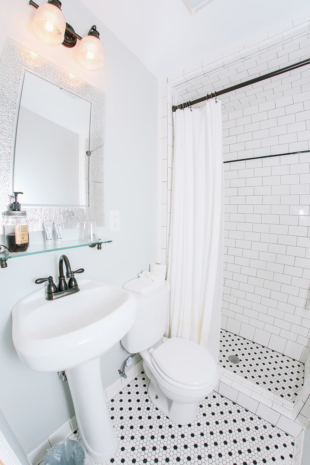 A bright, modern bathroom featuring a pedestal sink, toilet, and a shower with white tiles and a black-and-white patterned floor.