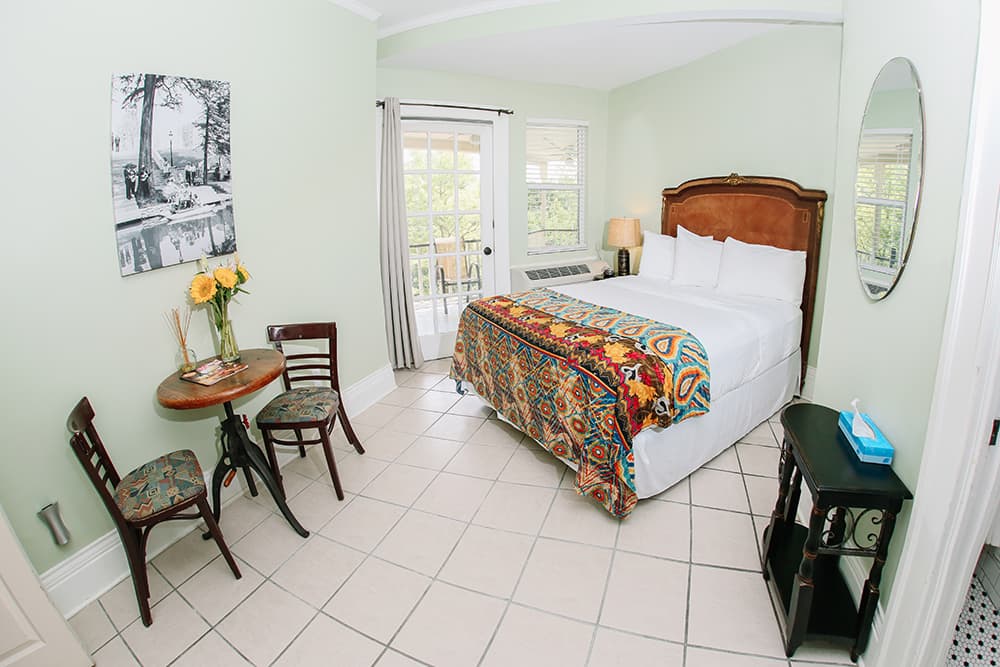 A cozy hotel room featuring a queen bed with a colorful patterned blanket, a small dining table with chairs, and large windows letting in natural light.