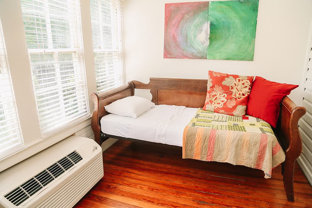 A cozy nook featuring a wooden daybed with colorful pillows, a quilt, and large windows allowing natural light.