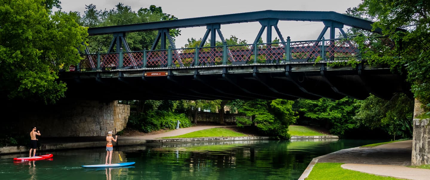 Two people paddleboarding under a historic metal bridge surrounded by lush greenery.