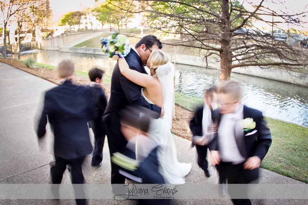 A couple embraces passionately in front of a river, surrounded by blurred figures of children in formal attire.