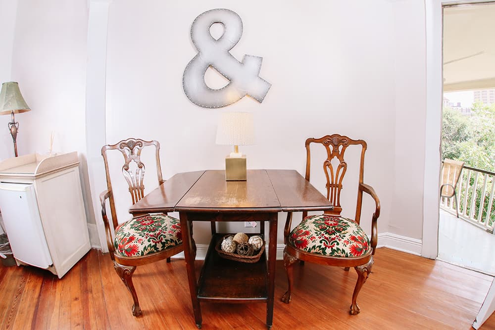 A small wooden table with two ornate chairs featuring floral cushions, accompanied by a lamp and an ampersand sign on the wall.