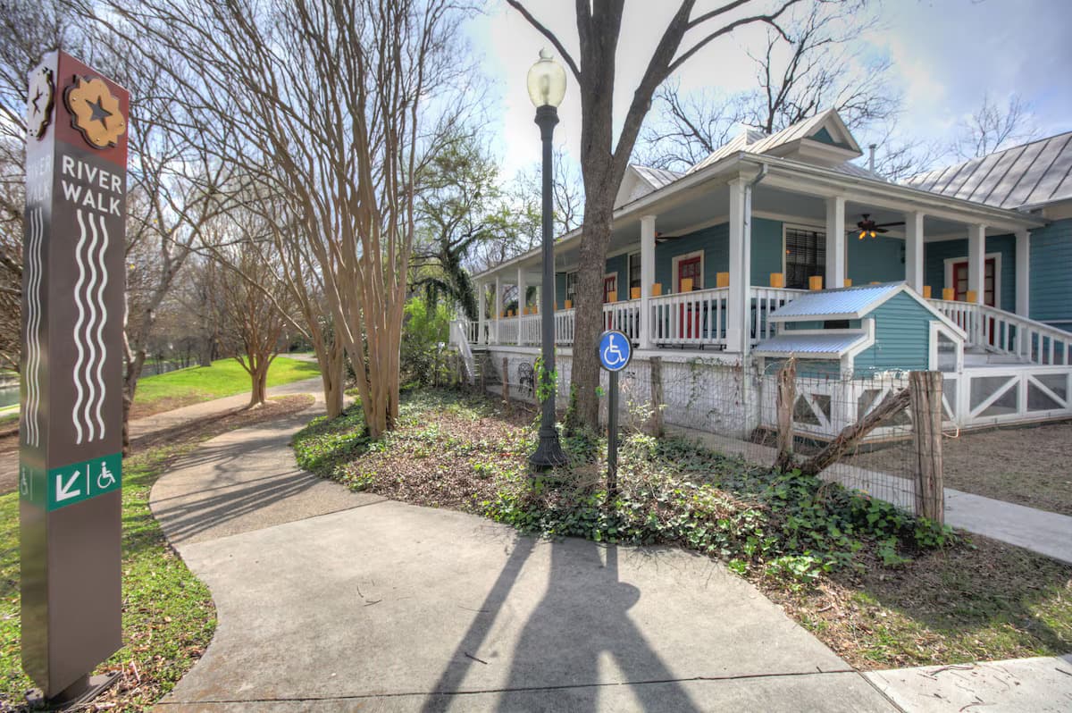A pathway leads to a blue house near the River Walk, featuring signage and greenery.