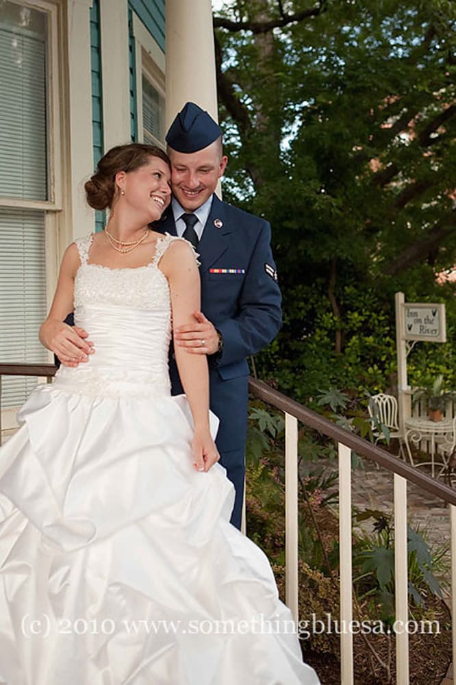 A bride in a white dress and an Air Force groom embrace joyfully on a porch.