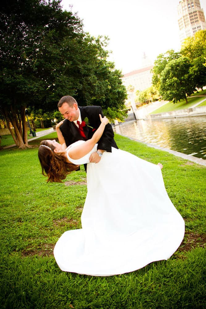 A groom dips his bride in a romantic pose near a pond surrounded by lush greenery.