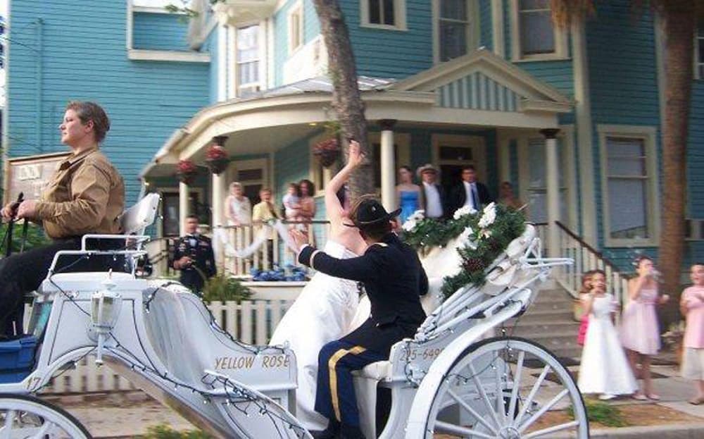 A bride waves from a decorated carriage as she is escorted by a groom in a formal uniform, surrounded by guests.