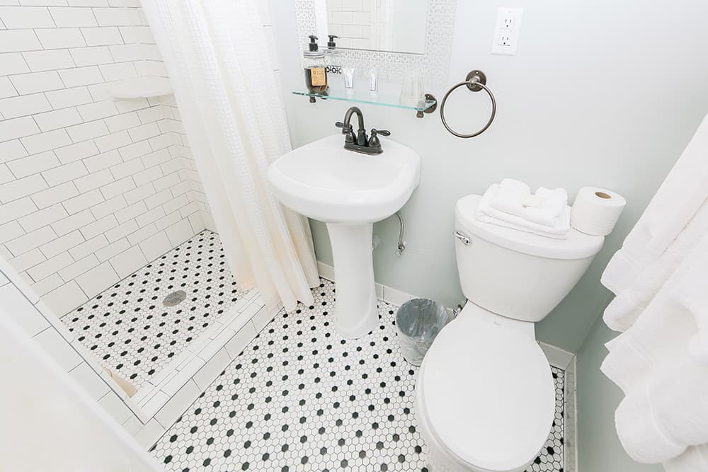 A modern bathroom featuring a shower, pedestal sink, and toilet with black and white patterned tile flooring.