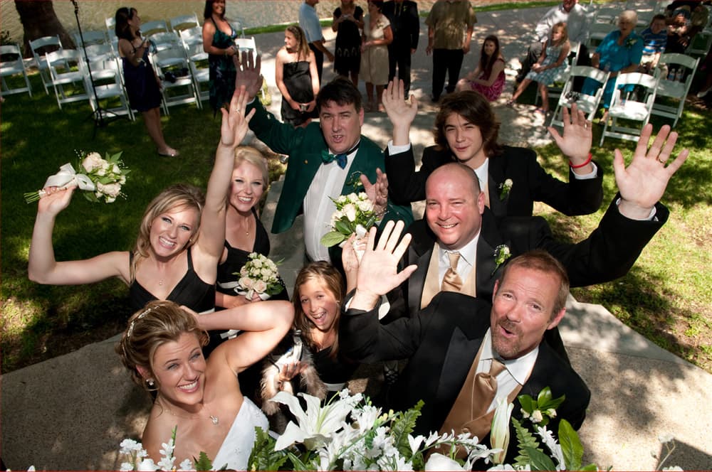 A joyful group of people in formal attire wave cheerfully at the camera during a wedding celebration.