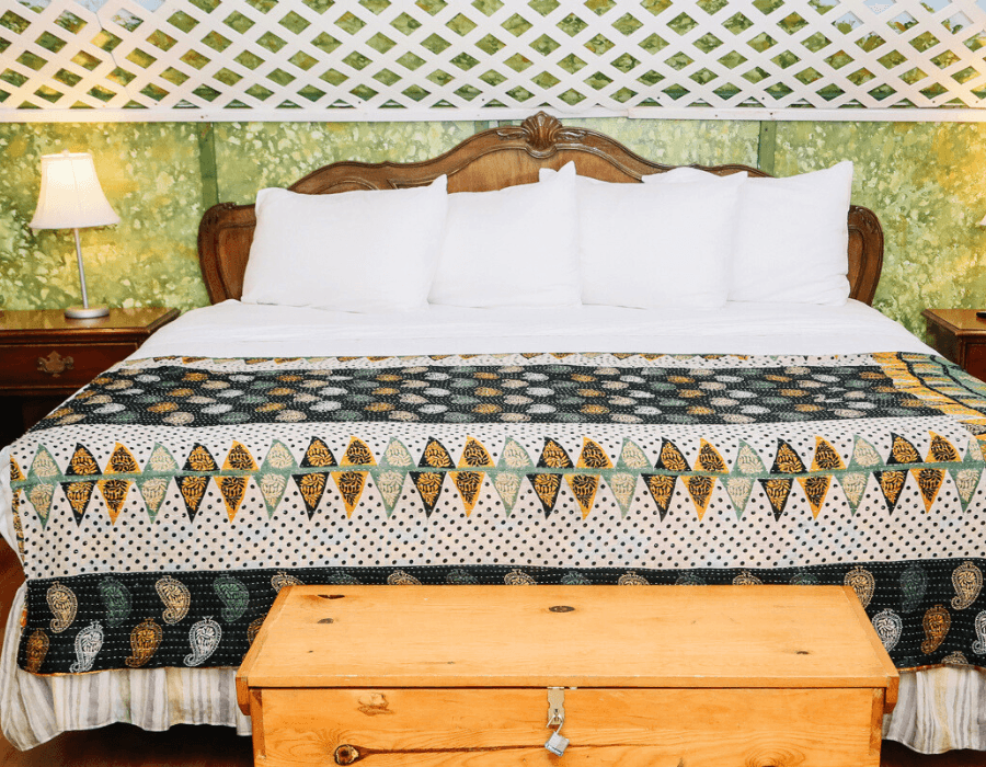 A neatly made bed with white pillows and a patterned quilt, accompanied by a wooden chest and a lamp in a cozy room.