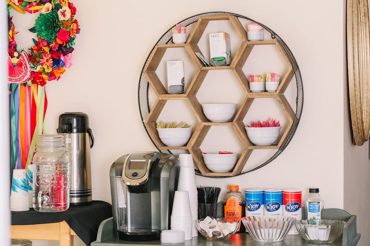 A decorative wall shelf holds various containers of snacks and drinks next to a coffee machine and water pitcher on a table.