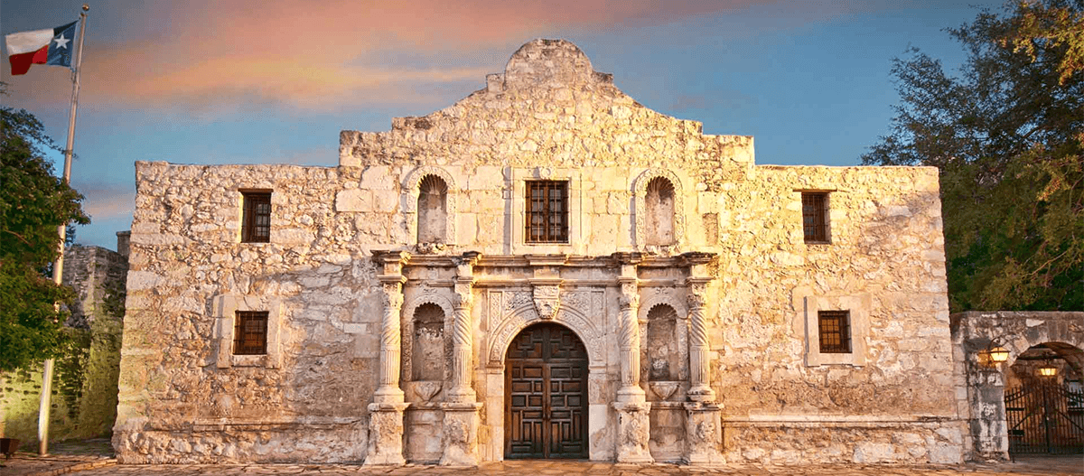 The Alamo, a historic stone structure in Texas, with a flag and trees in the background.