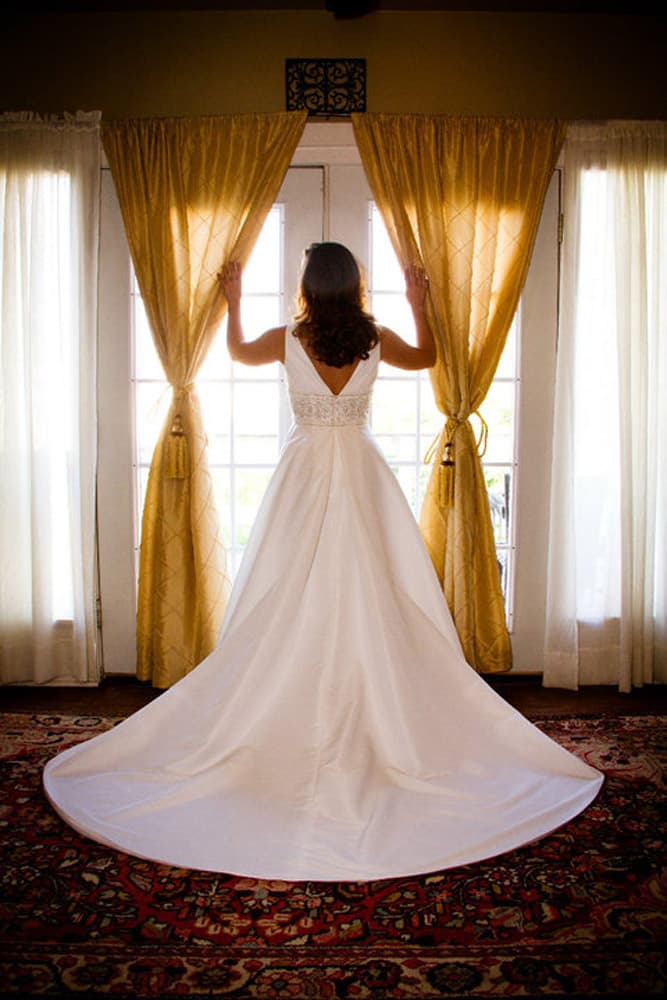 A bride in a white gown stands by a window, pulling back golden curtains.