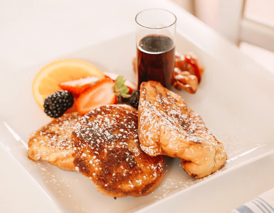 A plate of French toast topped with powdered sugar, accompanied by fresh fruit and a small glass of syrup.