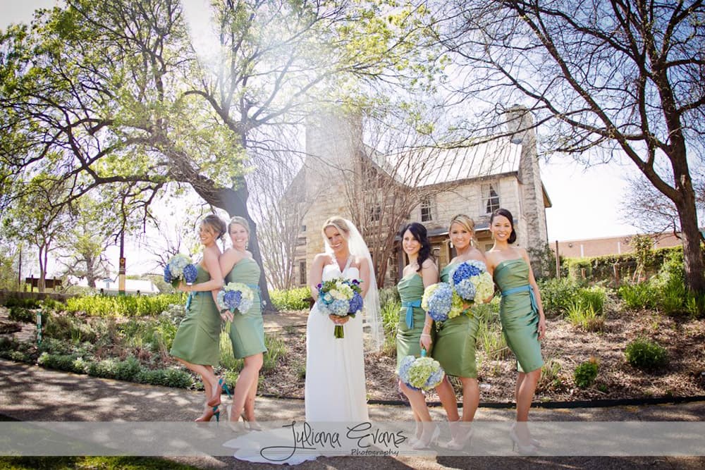 A bride in a white gown stands with six bridesmaids in green dresses, surrounded by lush greenery and a rustic building.