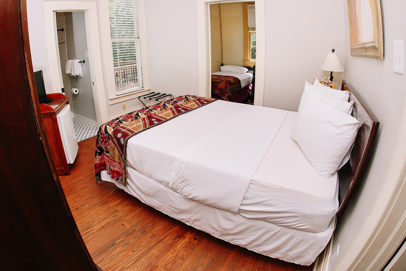 A cozy bedroom with a neatly made bed, wooden flooring, and a view into an adjoining room.