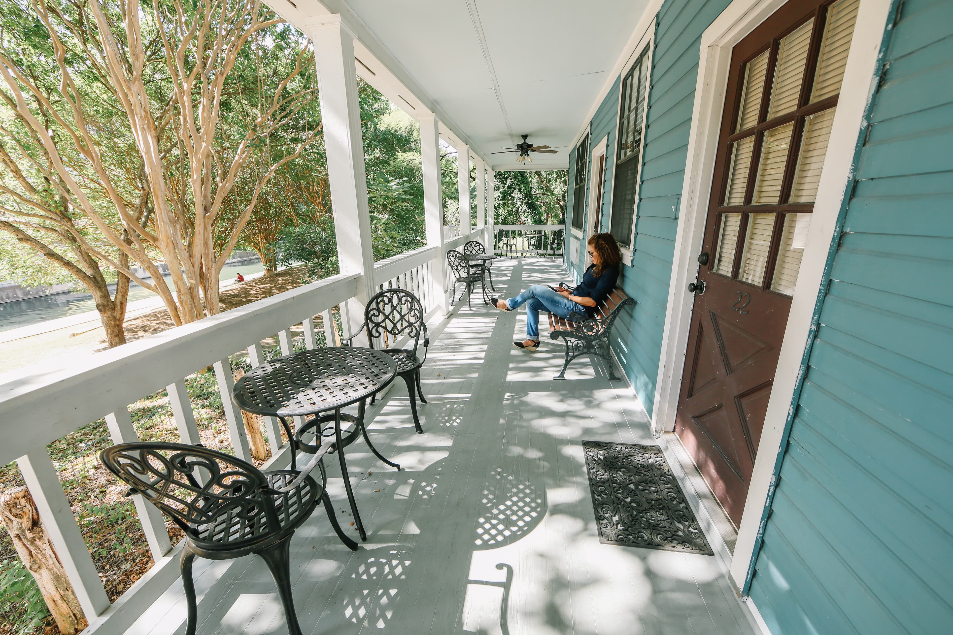 A woman is sitting on a porch, reading, with chairs and a door nearby.