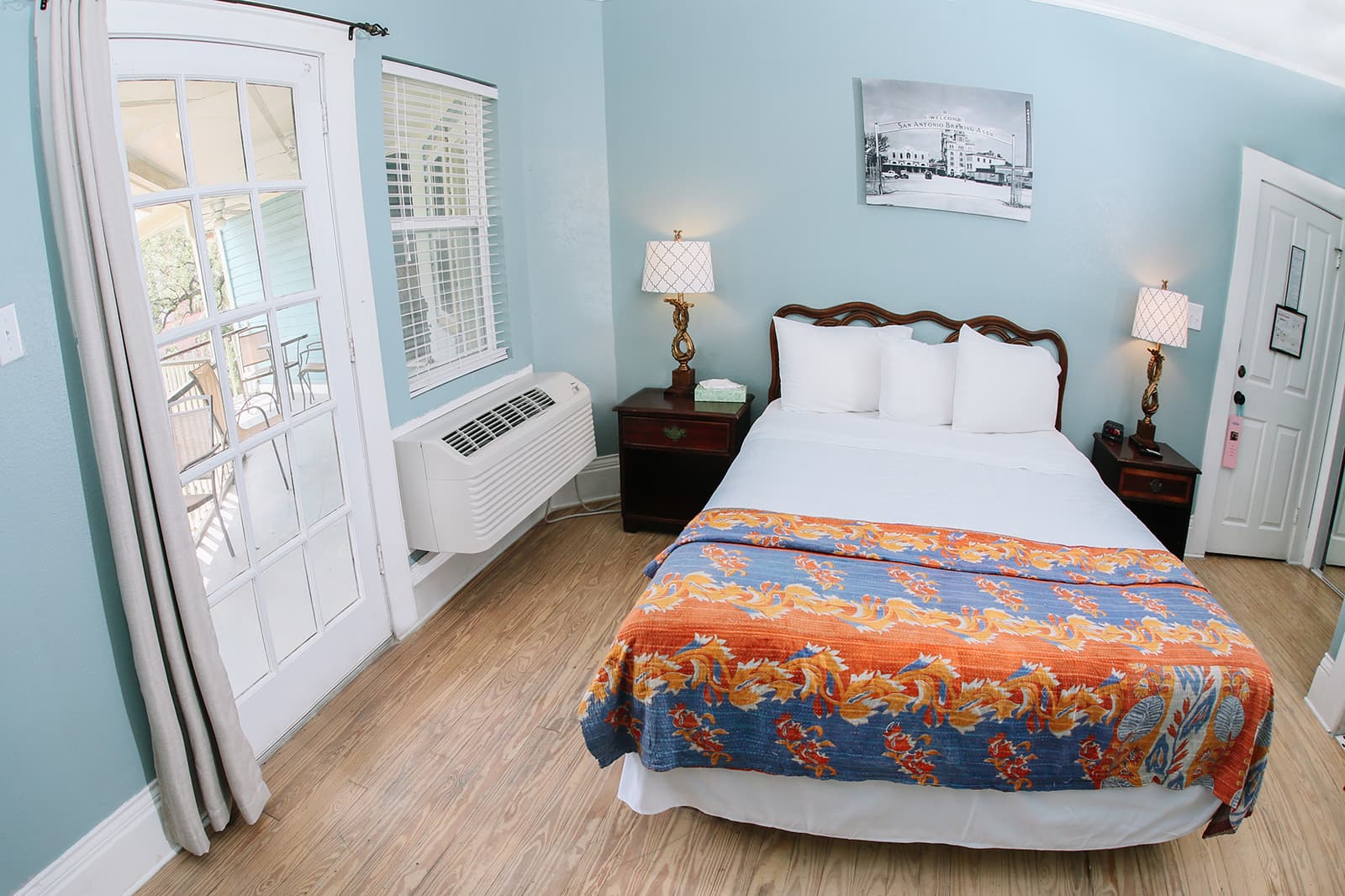 A cozy bedroom featuring a large bed with a colorful blanket, two bedside lamps, a window, and an air conditioning unit.