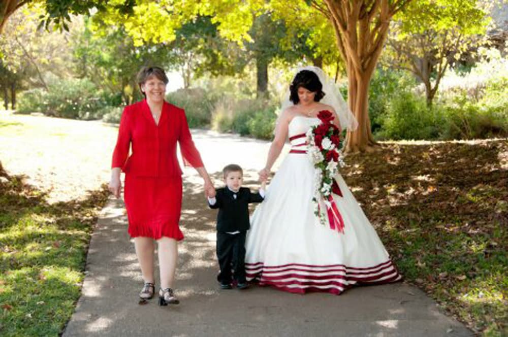 A bride in a white gown holds hands with a smiling child in a tuxedo, while walking alongside a woman in a red dress.