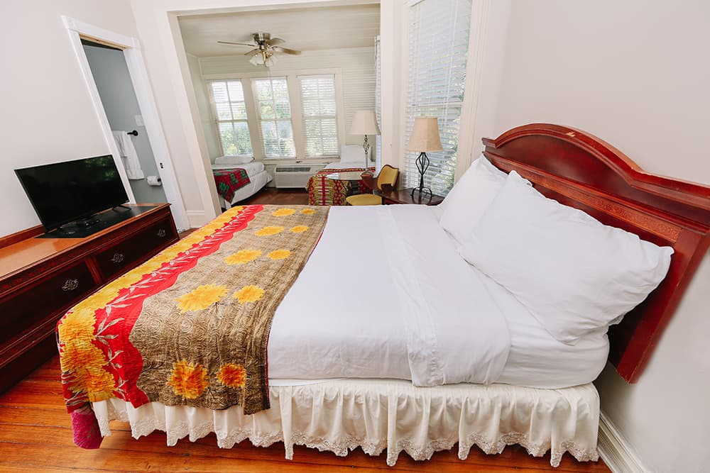 A cozy bedroom featuring a neatly made bed with colorful linens, a wooden dresser, and a view into a bright, airy sitting area.