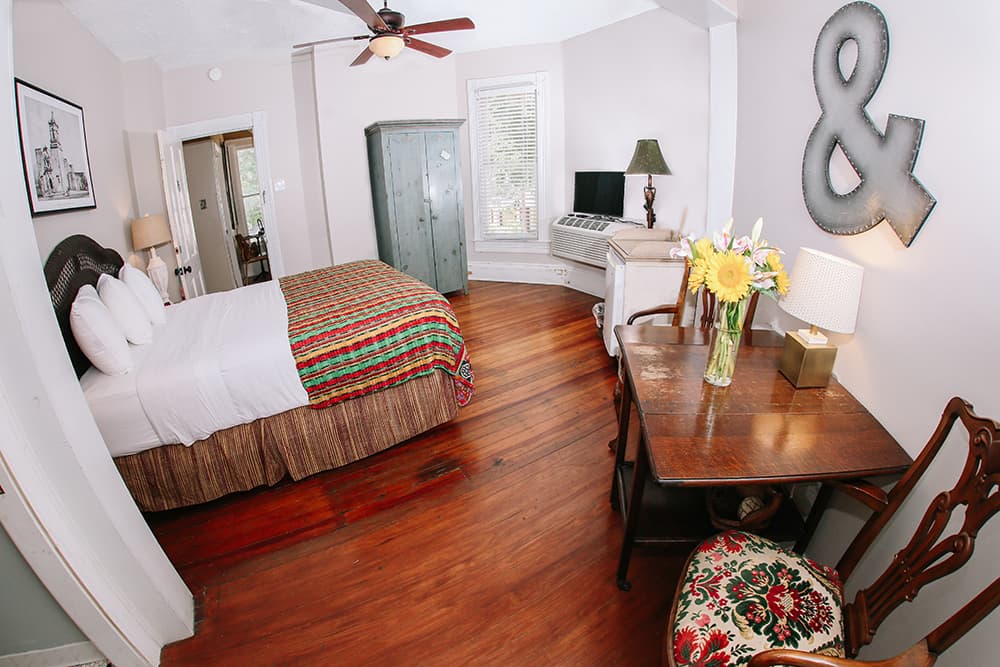Cozy bedroom featuring a bed with a colorful quilt, wooden flooring, a small workspace with flowers, and vintage furnishings.