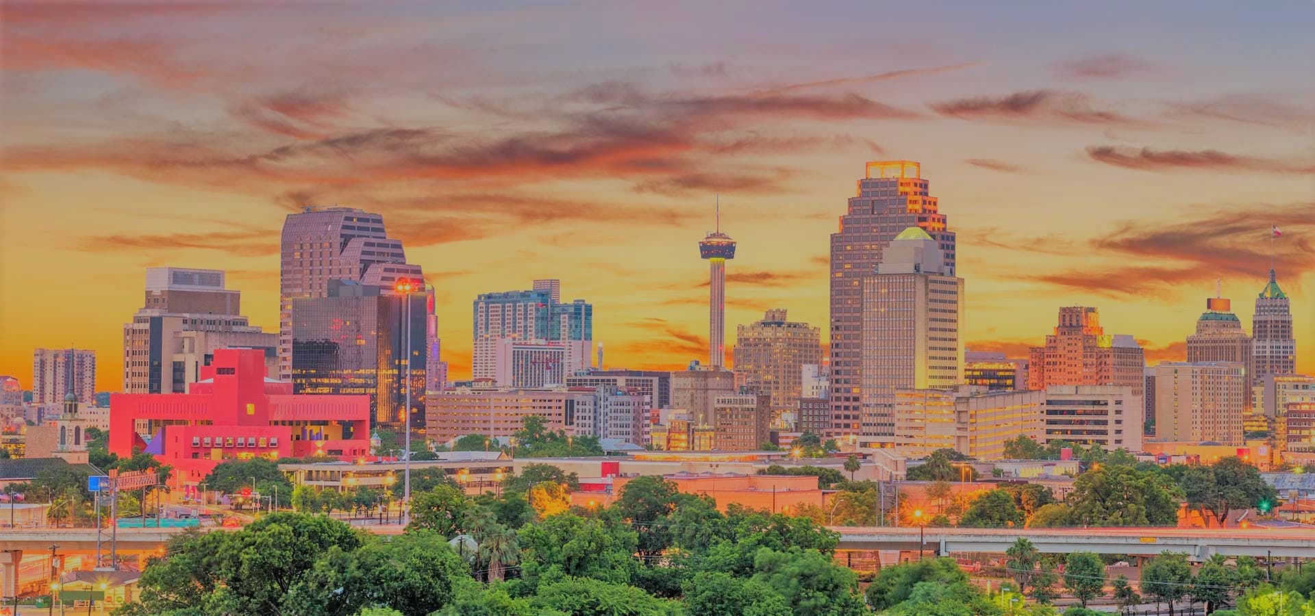 A vibrant sunset over the skyline of San Antonio, Texas, featuring diverse buildings and greenery in the foreground.