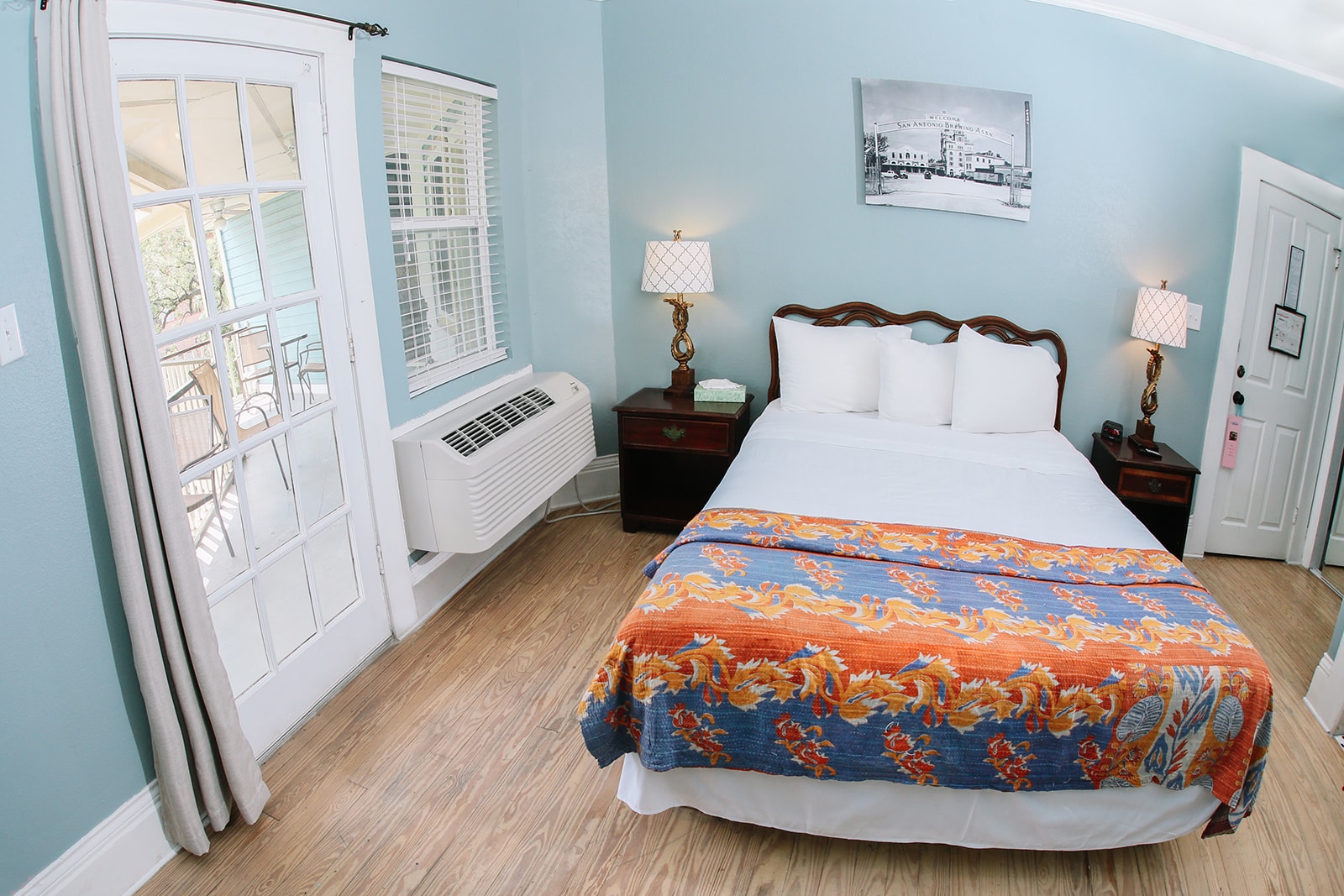 A cozy bedroom featuring a queen bed with colorful bedding, two lamps, and a windowed entrance.